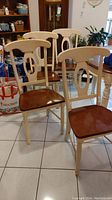 Photo showing four maple dining chairs with cream painted legs and backs, brown wood seats, arranged indoors on tile floor.