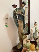 Tall Asian woman figurine standing beside a mirror, holding a rose and a fan. The figurine is elaborately dressed in traditional attire with detailed designs on the robe's sleeves and hem.
