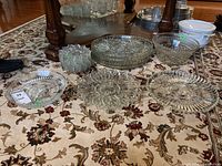 Photo showing many clear glass plates, bowls, and divided dishes arranged on a floral-pattern carpet under a wooden table
