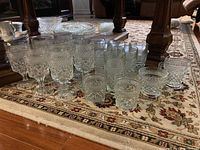 Overview of approximately 18 clear glass drinking pieces including stemmed glasses, tumblers, and other glass items placed on a patterned carpet.