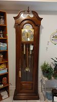 Full frontal view of tall wooden grandfather clock with arched top and glass door, showing entire clock standing against wall.