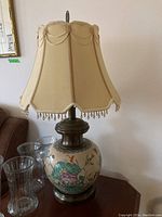 Full view of the lamp showing the ceramic base with floral and bird motifs, beige fabric shade with beaded fringe, finial on top.