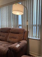 Tall brass colored floor lamp with a curved stand, featuring a white fabric-covered plastic cylindrical shade, positioned beside a brown upholstered loveseat.
