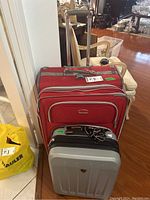 Photo showing large red Delsey suitcase positioned behind smaller silver-gray Heys suitcase, both with handles extended, set on a wooden floor near a white chair.