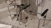 Two aluminum walkers of different sizes, several canes, shower chair, and cushions on a tiled bathroom floor