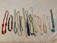Fourteen necklaces laid out flat on a white fabric background, showing variety in color, bead size, and chain style.