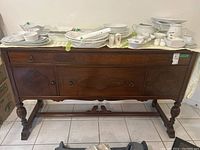Front view of wooden sideboard with various white ceramic dishes placed on top. Shows carved wood panel doors and drawers, metal pulls, and pedestal base.