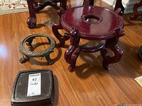 Three stands on wood floor: large round wooden stand with reddish finish, small square wooden stand, small round metal stand with decorative feet