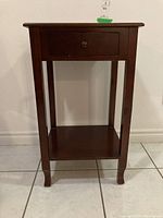 Front view of dark brown wooden side table with drawer and lower shelf, showing overall design and condition including scratches.