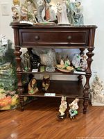 Front view of the wooden table showing drawer, shelves, turned legs, and items placed on top and on shelves.