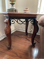 Wood side table with decorative carved legs and metal scrollwork support, placed next to a couch and window for size reference.