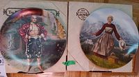 Photo of two collector plates from the set: The King and I plate showing a man in a red jacket and black pants, and The Sound of Music plate showing a woman with a goose and basket outdoors.