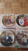 Photo shows the set of four "Annie" collector plates in their boxes, arranged in a 2x2 layout on a wooden floor.