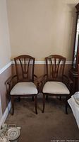 Two wooden arm chairs with upholstered light fabric seats and decorative back design in corner of room