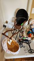 Photo showing the vintage wicker doll carriage with black fabric canopy, oval lace-trimmed basket, black metal tricycle, and small riding horse doll accessory on a table.