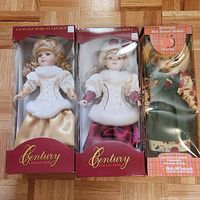 Front view of three boxed porcelain dolls: two from Century Collection with maroon boxes and one Ann of Green Gables doll in an orange box.