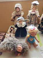 Group photo showing a variety of dolls including porcelain and mixed material dolls arranged on a surface with a box and papers beneath.