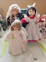 Four porcelain dolls, including Trish Romance Patient Angel doll in pink dress with certificate and box, two dolls on stands with floral and dark green dresses, and one small doll in pink dress.