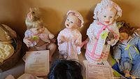 Three porcelain dolls positioned side by side with their certificates of authenticity visible in the foreground. Dolls dressed in pastel colors including pink and white, with detailed facial features and clothing.