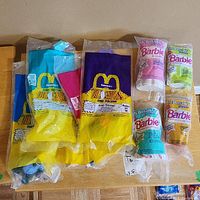 Photo of six Dino-Motion dinosaur toys in sealed bags with McDonald's branding on a wooden surface beside five Barbie styling dolls in their packaging.