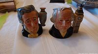 Two Royal Doulton Toby Jugs depicting Sir Henry and John Doulton on a table.