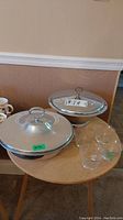 Photo of two metal lidded serving dishes with glass inserts and four clear glass nappies on small table.