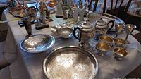 Full overview of all silver plated items including teapot, coffee pot, creamers, sugar bowls, cups, and serving trays on a table with other decorative pieces in background
