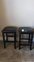 Two matching wood stools with black padded seats placed side by side on carpeted floor against beige wall.