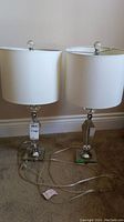 Front view of two identical table lamps with white fabric shades and clear plastic and metal bases placed on carpet.