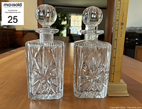 Pair of square-shaped pressed glass decanters with stoppers sitting side-by-side on a wooden table, ruler showing height of 9.5 inches.
