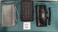 Three clutch purses laid out on teal fabric: silver rhinestone clutch with wrist strap on left, vintage embroidered black fabric clutch in center, and black Kenneth Cole wristlet clutch on right.