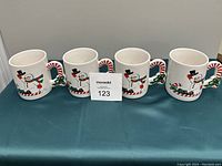 Front view of four white ceramic Christmas mugs with snowman decorations and candy cane shaped handles on a green cloth and white backdrop, all in excellent condition.