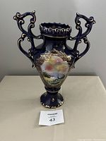 Front view of the blue Alcobaca vase showing painted pastoral scene with trees and water, gold detailing on handles and base.