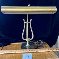 Front view of brass piano lamp showing overall design, lamp head, lyre shaped stand, base, power cord, and ruler for size reference.