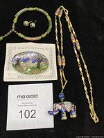 Photo showing the cloisonne jewelry lot including a long gold-tone metal chain necklace with multicolor cloisonne elephant pendant, green cloisonne bracelet, matching green post earrings, and a hand-painted outdoor scene pin.
