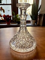 Front view of vintage crystal cut glass decanter with stopper on wooden table near window with plants visible in background.