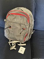 Front view of grey Eastport North backpack with red zippers and two elastic cords on front compartment, shown against a blue background