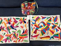 Two colorful appliqued fabric wall hangings with bright animal and floral designs, and a small purse with similar appliqued decorative motifs placed against dark background.