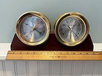 Frontal view of the barometer and clock set on a wooden base alongside a ruler showing the base width as approximately 13 inches.