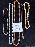 Photo showing five necklaces laid out flat with a MaxSold label in the center. Necklaces include a long 26 inch wooden bead necklace with gold metal beads, a 16 inch light wooden stretch bead necklace, a 24 inch sage/white/grey stone bead necklace, a 15 inch iridescent small bead necklace, and an 18 inch gold metal chain with a stone pendant.