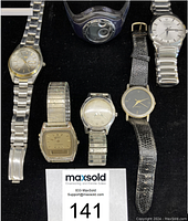 Photo showing six men's wristwatches laid out on a black surface including various styles with metal and leather bands.