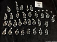 Photo showing 34 clear faceted chandelier crystals arranged on black fabric from large to small, mostly teardrop shapes with metal hooks attached for hanging.