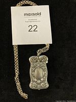 Close-up photo of vintage late 19th-century silver match case pendant with ornate floral embossed design and attached heavy silver chain, showing a tag labeled '22'.