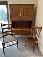 Front view of oak Hoosier cabinet with two wooden chairs beside it in a carpeted room near window, showing cabinet doors, drawers and chair details