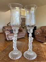 Pair of Baccarat crystal cherub putti candlesticks on wooden table with living room background, showing entire candlesticks with frosted cherub stems and etched hurricane shades.