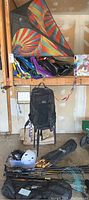 Wide view showing two large vibrantly colored kites, folded kites, a black backpack hanging, helmets, hiking poles, and other gear on shelves and floor.