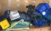 Camping gear including blue Columbia tent bag with owner's manual, yellow Coleman lantern, black metal tripod, blue water container, and rolled sleeping pads.