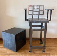 Front angle view of the handmade steel chair and solid steel cube placed on wood floor against a white wall.
