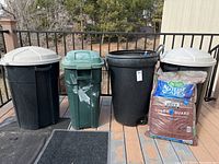 Front view of three trash cans (two black with dome lids, one green) and a bag of mulch on patio floor