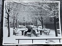 Black and white photo of person sitting on snow-covered benches holding umbrella under leafless trees